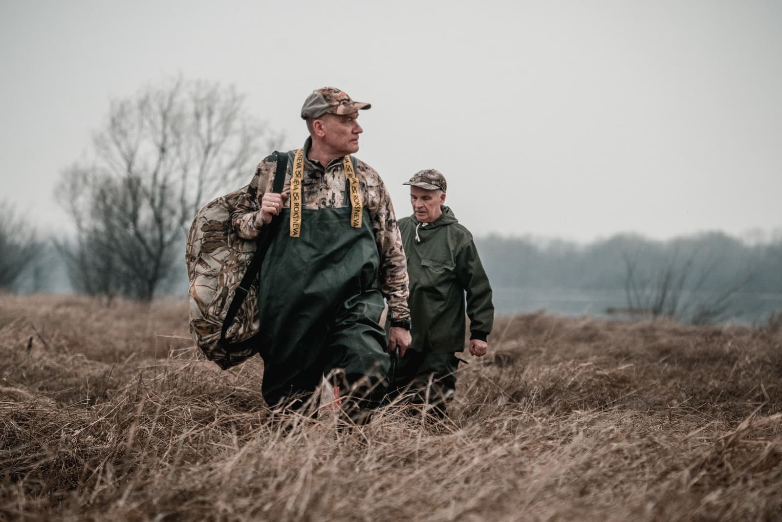 Two men in green chest waders and camo walking through dry marsh grass